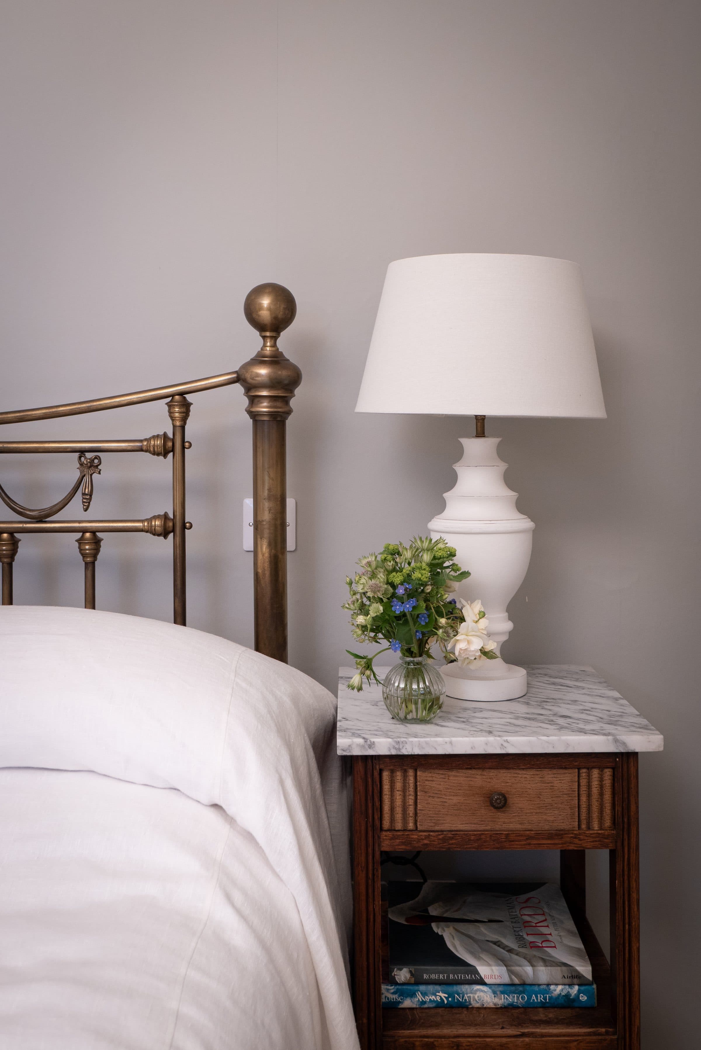 A brass bedstead with white linen, a marble-topped bedside table, lamp and a small vase of flowers.