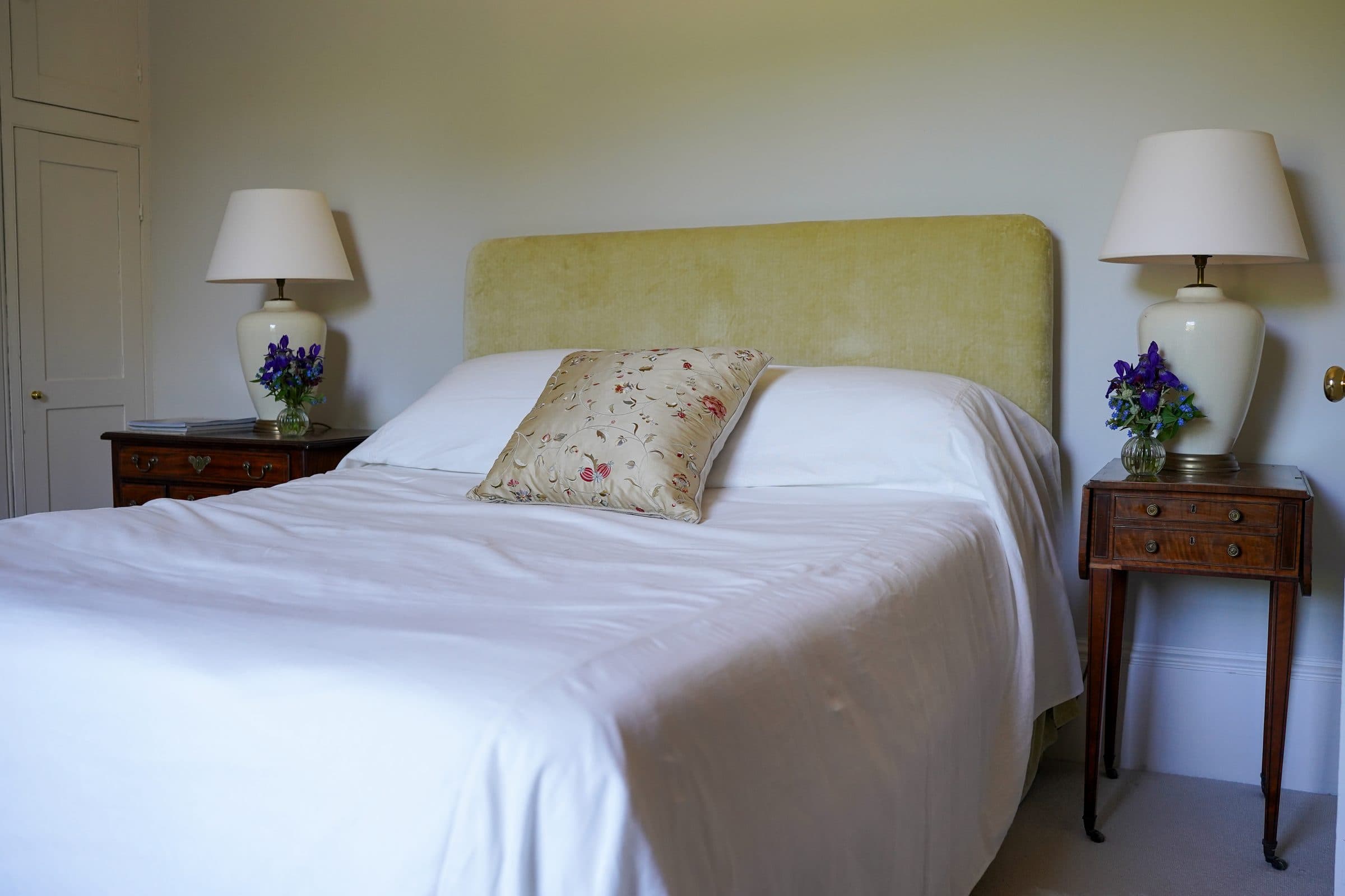 A double bed with a green upholstered headboard between matching lamps and antique bedside tables.