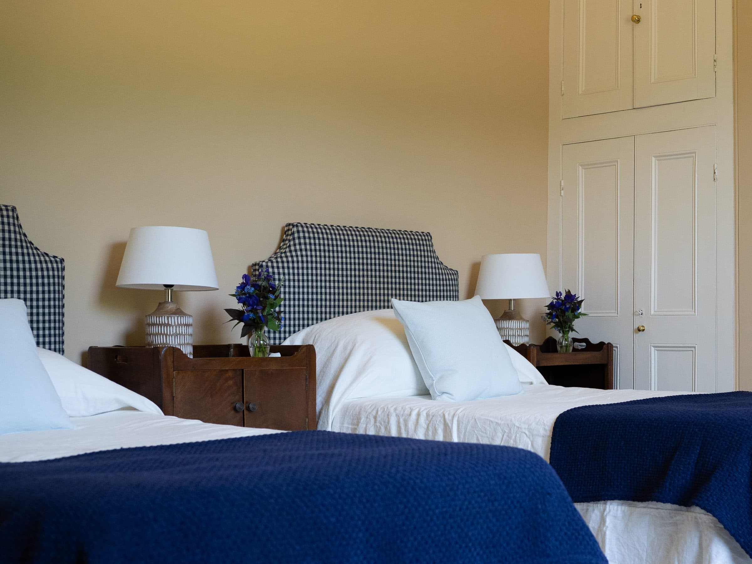 Twin beds with checked headboards and blue blankets, a lamp and flowers on the table between them.