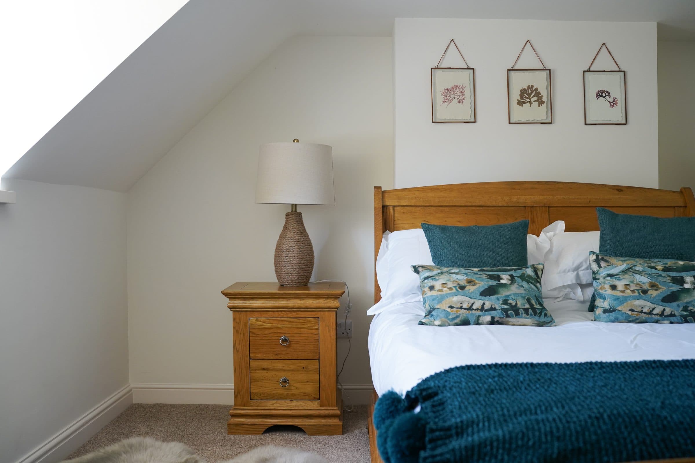 A Cottage bedroom under the eaves, with an oak bed, teal blanket and prints above the headboard.