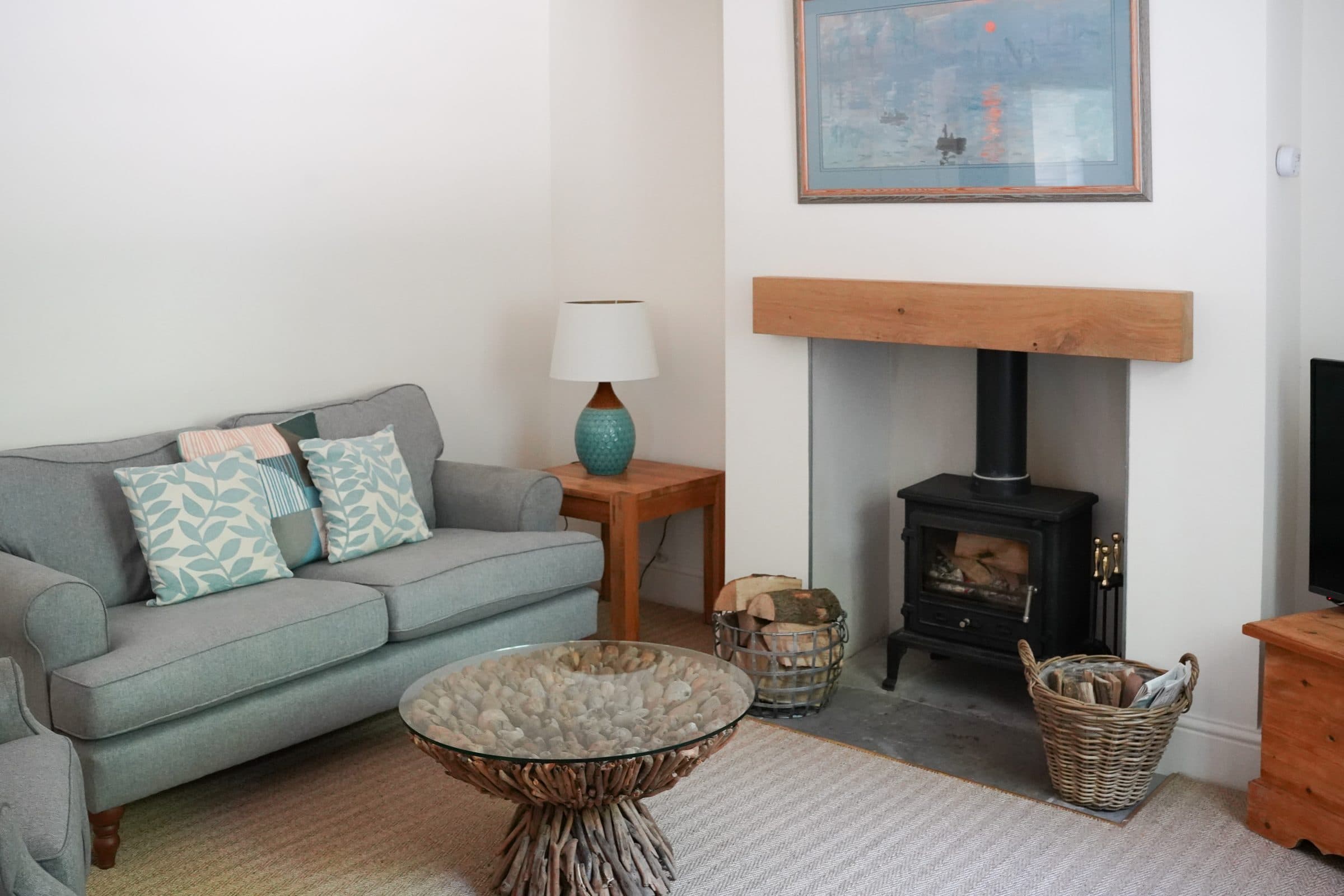 The Cottage sitting room with a wood burner, soft green sofa and a basket of logs beside the hearth.