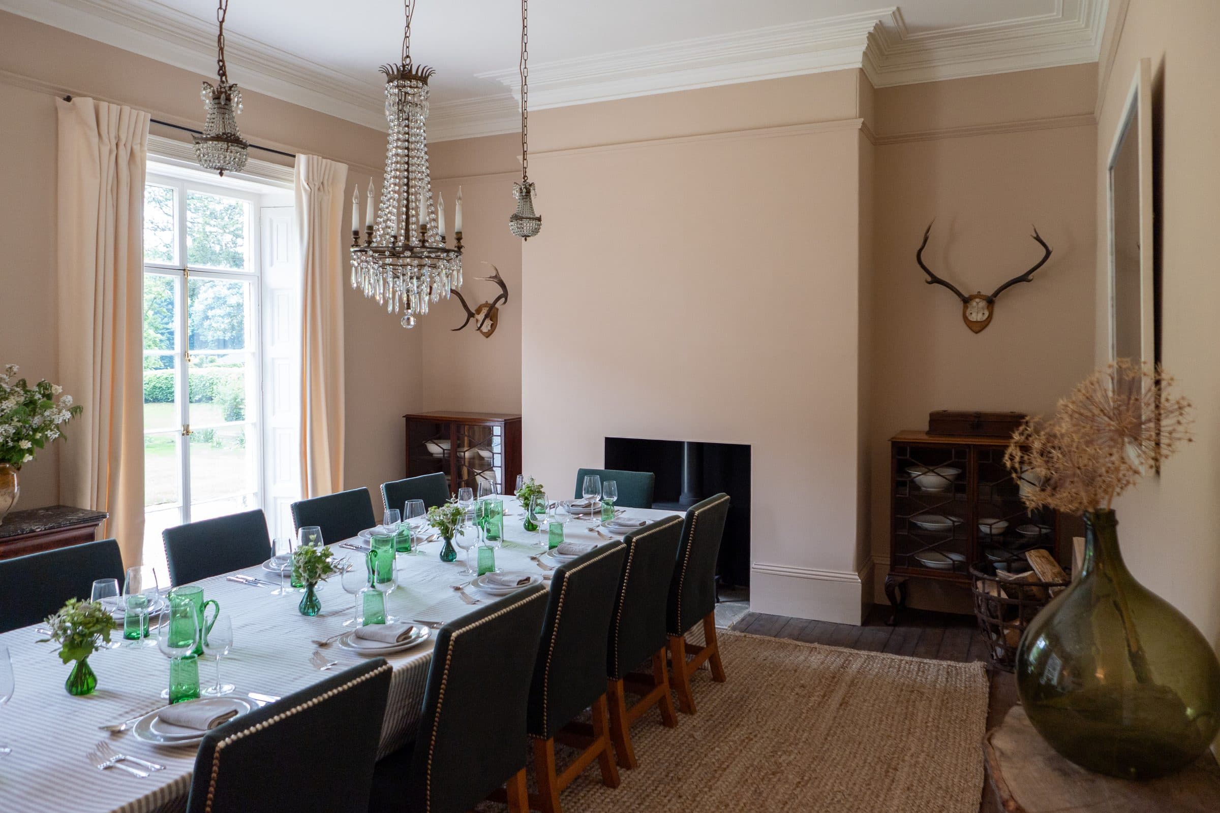 The dining room laid for sixteen with green glassware and white linen, antlers on the wall and a glazed cabinet at the end.