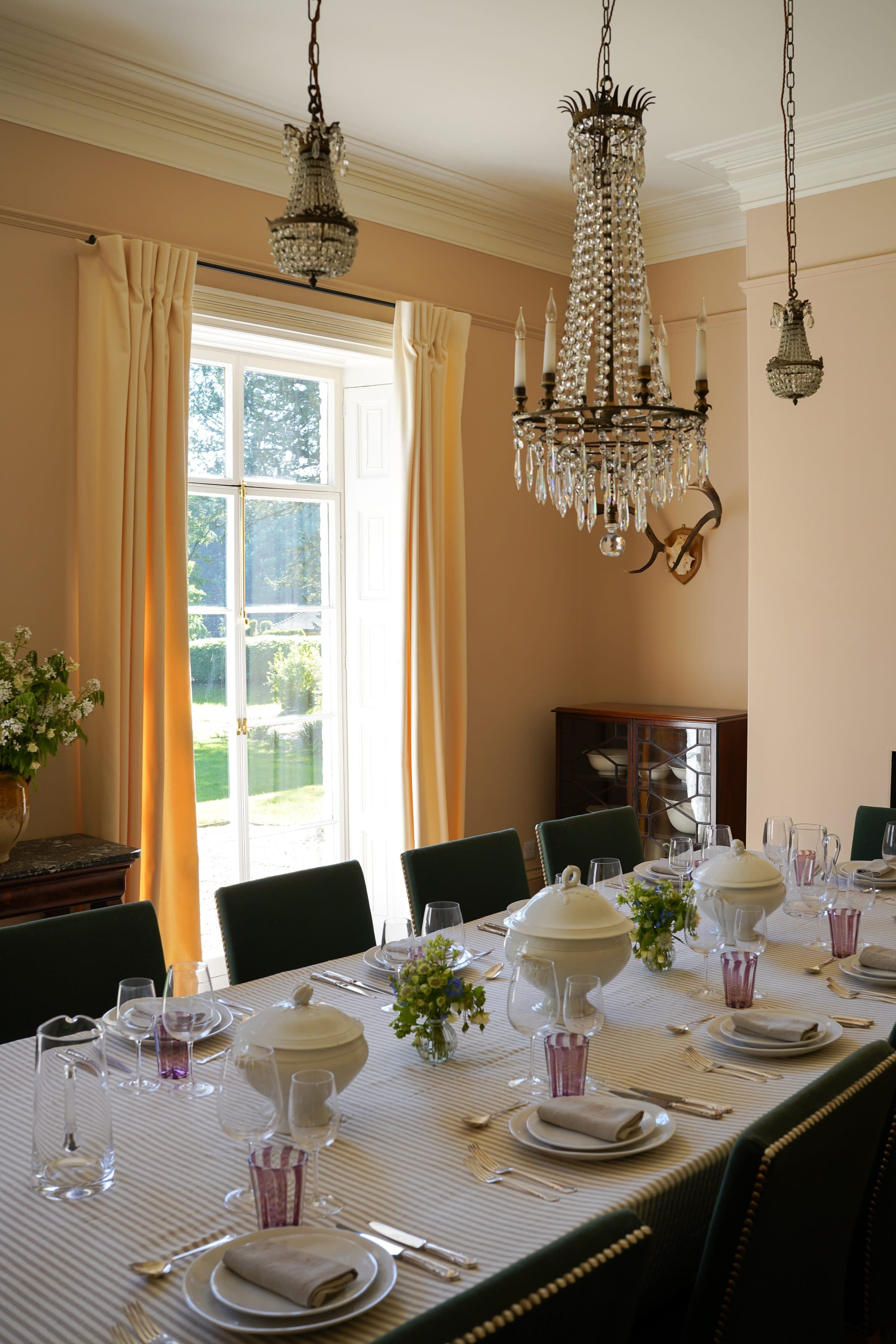 The dining room laid for a long lunch with amethyst glasses and striped linen, white tureens and posies down the table beneath the crystal chandeliers, the garden door standing open.