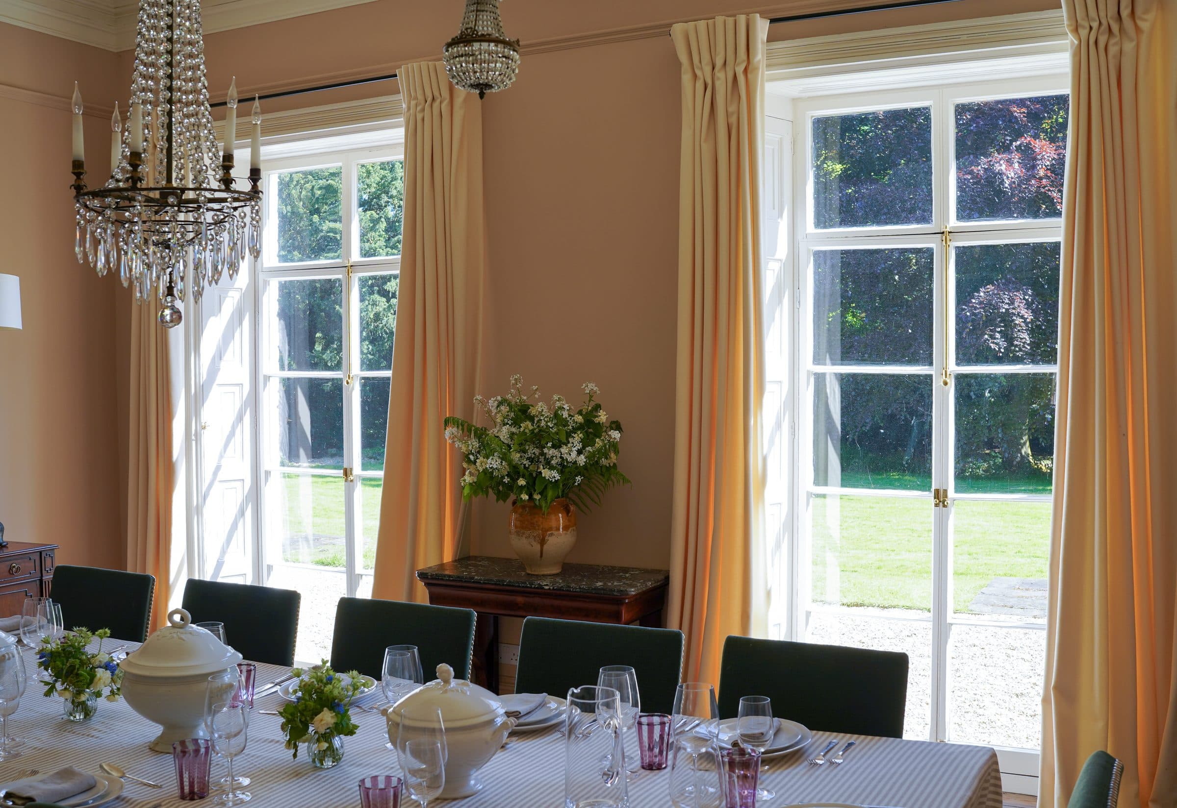 The full dining room set for a long lunch beneath two crystal chandeliers, with garden light from two tall windows.