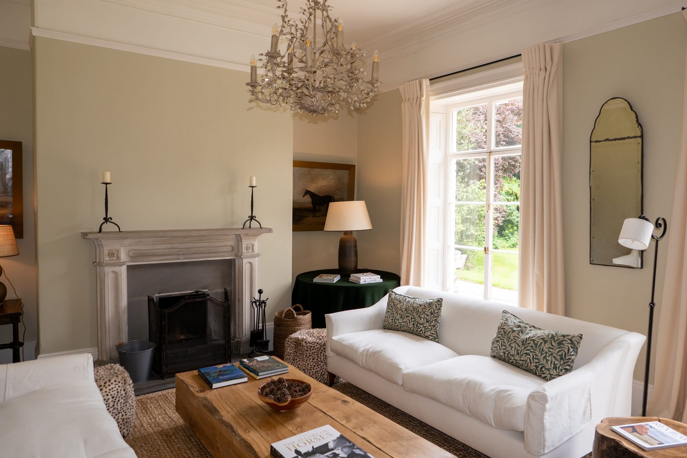 The drawing room with white sofas either side of the stone fireplace, a gilt chandelier above and the garden through tall sash windows.